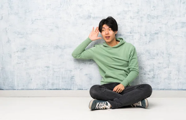 耳に手を置いて何かを聞いて床に座っているアジア人男性 — ストック写真