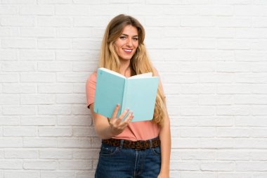 Beyaz tuğlalı duvarın üzerinde elinde kitap tutan ve okuyan genç sarışın kadın.