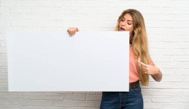 Beyaz tuğlalı duvarın üzerinden geçen genç sarışın kadın bir konsept eklemek için boş beyaz bir levha tutuyor.