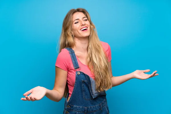 Young blonde woman with overalls over isolated blue wall smiling