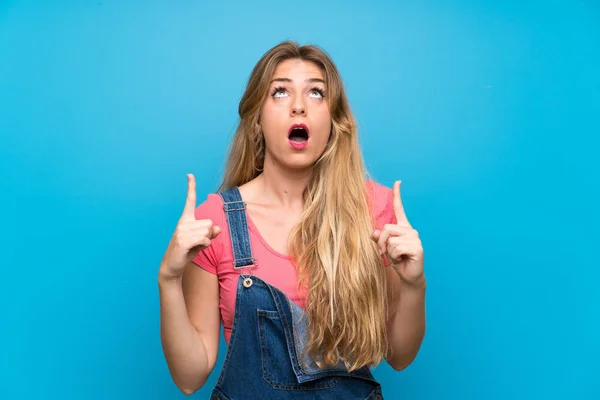 Young Blonde Woman Overalls Isolated Blue Wall Surprised Pointing — ストック写真