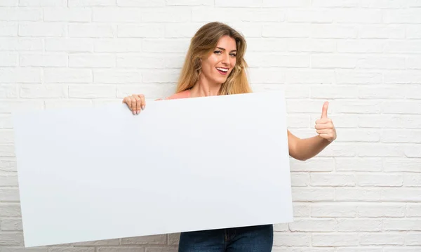白砖墙上的金发年轻女子拿着一个空的白色标语牌插入一个概念 — 图库照片
