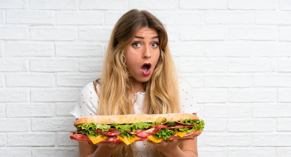 Joven mujer rubia sosteniendo un gran sándwich haciendo gesto sorpresa —  Fotos de Stock