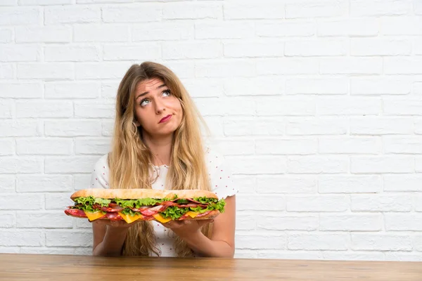 Young blonde woman holding a big sandwich having doubts — Stock Photo, Image