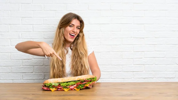 Szczęśliwy młody Blondynka kobieta trzyma a duży Sandwich — Zdjęcie stockowe
