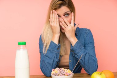 Bir kase tahıl gevreğiyle gözlerini kapayan ve parmaklarından içeri bakan genç sarışın kadın.