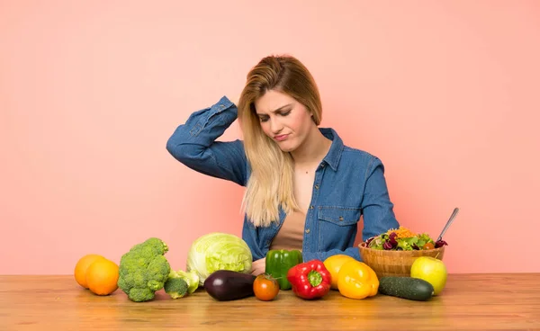 Young blonde woman with many vegetables with an expression of frustration and not understanding