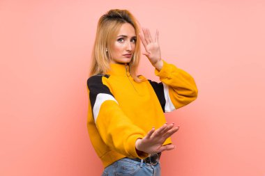 Soyutlanmış pembe arka planda genç sarışın kadın gergin ellerini öne uzatıyor.