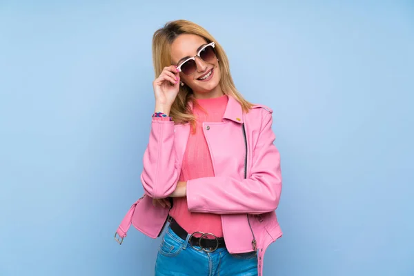 Молодая Блондинка Розовой Куртке Изолированном Голубом Фоне Очками Счастливой — стоковое фото