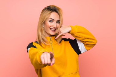 Soyutlanmış pembe arka planda genç sarışın bir kadın telefon hareketi yapıyor ve ön tarafı işaret ediyor.