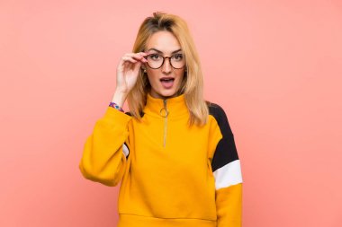 Gözlüklü ve şaşırmış, pembe arka planı olan genç sarışın bir kadın.