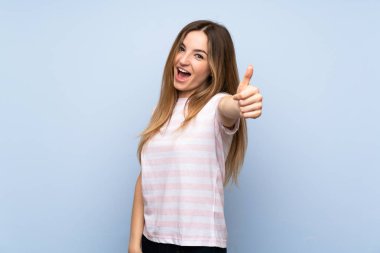 Soyutlanmış mavi arka planı olan genç bir kadın. Başparmakları havada çünkü iyi bir şey oldu.