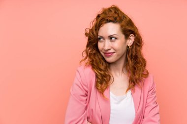 Izole pembe duvar ayakta üzerinde takım elbiseli Redhead kadın ayakta ve yan bakıyor