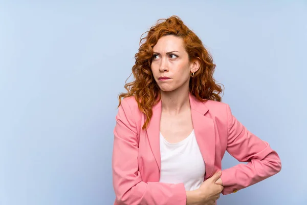 Redhead woman in suit over isolated blue wall with confuse face expression