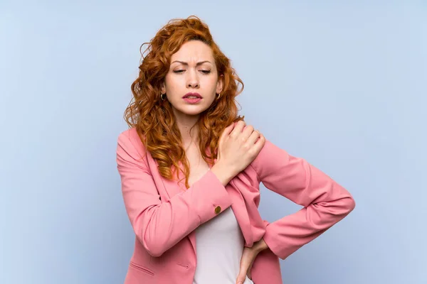 Redhead woman in suit over isolated blue wall suffering from pain in shoulder for having made an effort