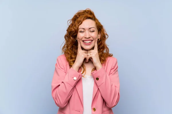 Redhead woman in suit over isolated blue wall smiling with a happy and pleasant expression