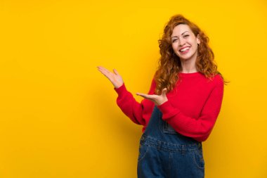 Izole sarı duvar üzerinde tulum ile Redhead kadın gelmeye davet için yan ellerini uzatıyor