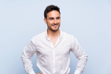 Izole mavi arka plan üzerinde yakışıklı genç adam kalça kolları ile poz ve gülümseyerek