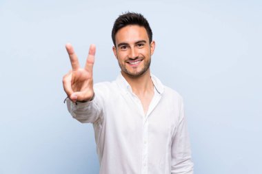 Izole mavi arka plan üzerinde Yakışıklı genç adam gülümseyerek ve zafer işareti gösteren