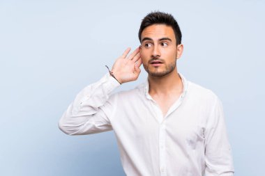 İzole mavi arka plan üzerinde yakışıklı genç adam kulak el koyarak bir şey dinliyor