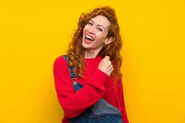 Redhead woman with overalls over isolated yellow wall celebrating a victory