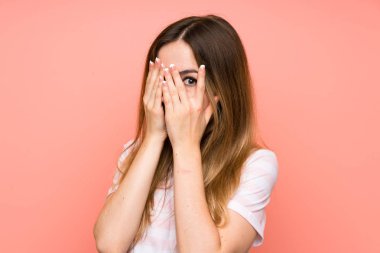 İzole edilmiş pembe duvarın üzerinde genç bir kadın gözlerini kapatıyor ve parmaklarından bakıyor.