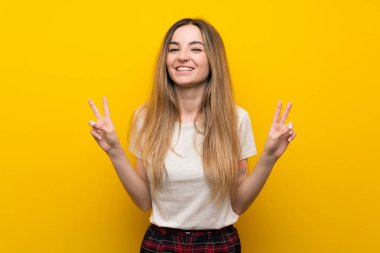 İzole edilmiş sarı duvar üzerinde genç bir kadın zafer jesti yapıyor.