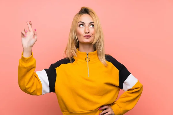 Young blonde woman over isolated pink background with fingers crossing and wishing the best