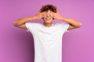İzole edilmiş mor duvarın üzerinde gözlerini elleriyle kapayan genç bir Afrikalı Amerikalı.