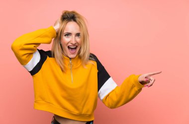 Soyutlanmış pembe arka planı olan genç sarışın kadın şaşırdı ve işaret etti.