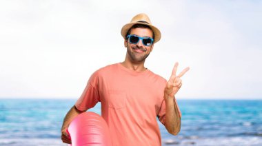 Yaz tatili gülümseyen ve plajda neşeli bir yüz ile zafer işareti gösteren üzerinde şapka ve güneş gözlüğü ile adam