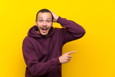 Sarı duvar üzerinde Sweatshirt ile Kolombiyalı adam sürpriz ve tarafına parmak işaret
