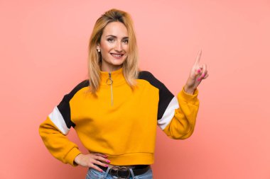 Soyutlanmış pembe arka planı olan genç sarışın kadın en iyinin işareti olarak parmağını kaldırıyor ve gösteriyor.