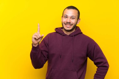 Sarı duvara gösteren ve en iyi işareti bir parmak kaldırma üzerinde Sweatshirt ile Kolombiyalı adam