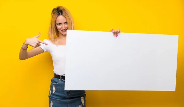 年轻的金发女子翻过孤立的黄墙 拿着空白的白色标语牌插入一个概念 — 图库照片