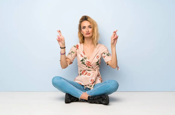 Young blonde woman sitting on the floor with fingers crossing and wishing the best