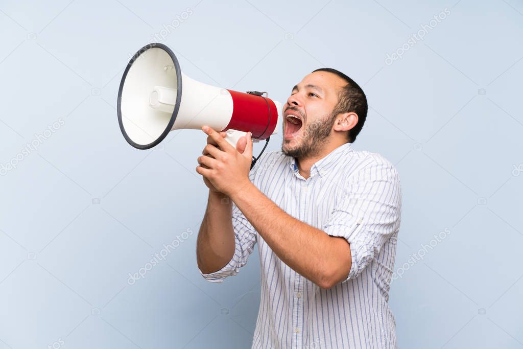 Colombian man over isolated blue wall shouting through a megaphone