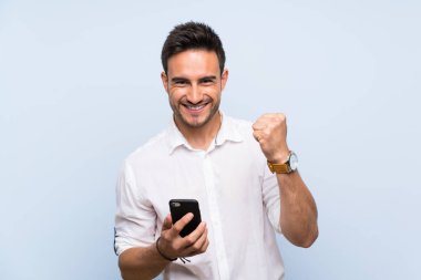 Zafer pozisyonunda telefon ile izole mavi arka plan üzerinde yakışıklı genç adam