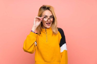 Gözlüklü ve şaşırmış, pembe arka planı olan genç sarışın bir kadın.