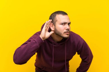 Sarı duvar üzerinde Sweatshirt ile Kolombiyalı adam kulak el koyarak bir şey dinliyor