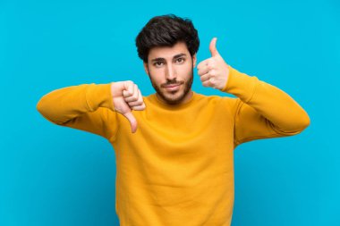 Izole mavi duvar üzerinde yakışıklı iyi-kötü işareti yapıyor. Evet ya da değil arasında kararsız