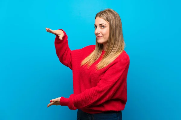 Woman with red sweater over blue wall holding copyspace to insert an ad