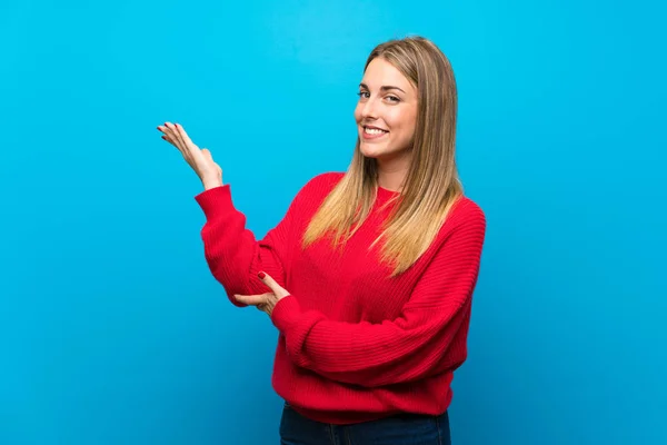 Woman with red sweater over blue wall extending hands to the side for inviting to come