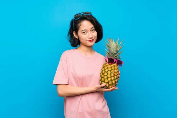 Asiatisk ung kvinna över isolerad blå bakgrund innehar en ananas med sol glasögon — Stockfoto