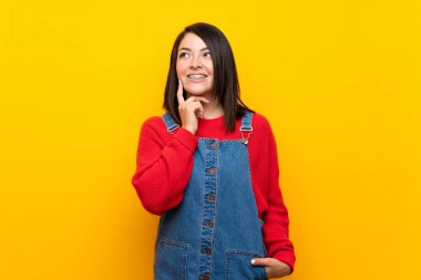 Young Mexican woman with overalls over yellow wall thinking an idea while looking up