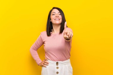 Young Mexican woman over isolated yellow background showing and lifting a finger