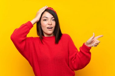 Young Mexican woman with red sweater over yellow wall surprised and pointing finger to the side