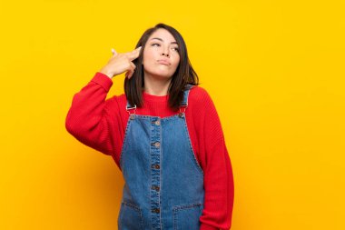 Young Mexican woman with overalls over yellow wall with problems making suicide gesture
