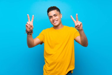 Young man with yellow shirt over isolated blue background showing victory sign with both hands