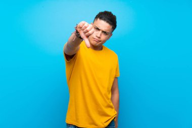 Young man with yellow shirt over isolated blue background showing thumb down with negative expression